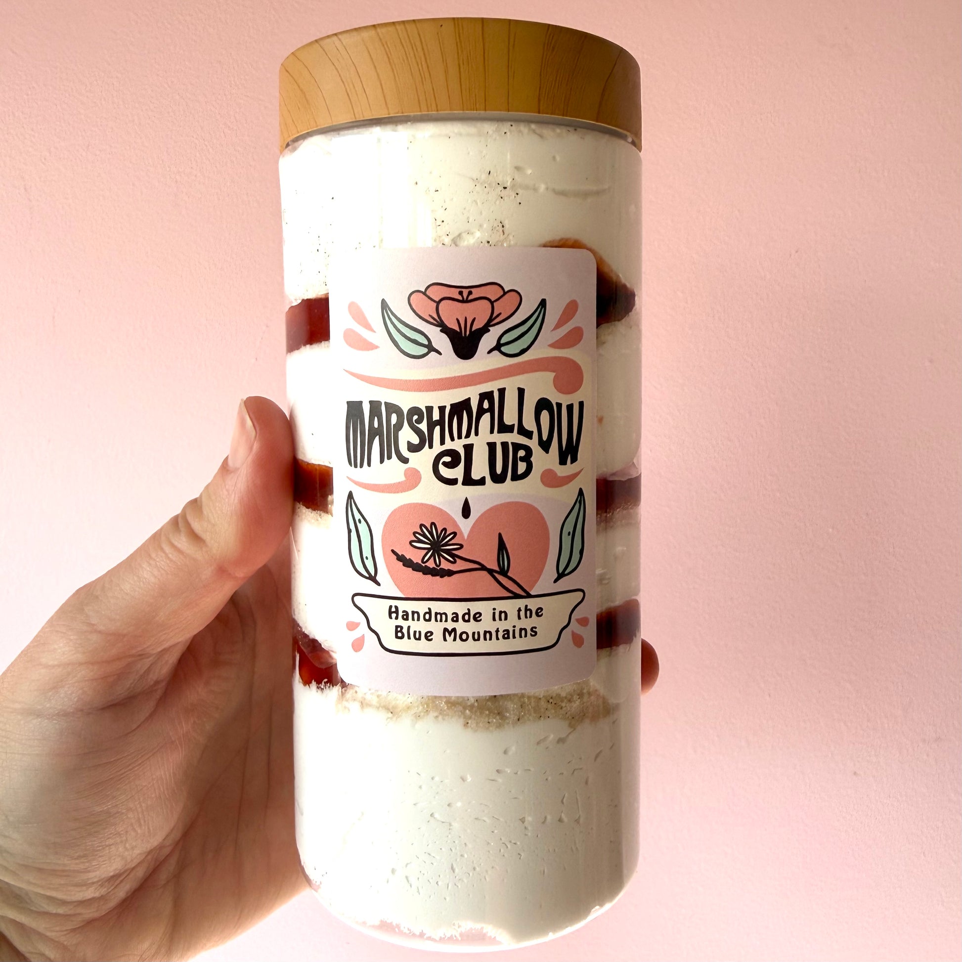 A hand holding a clear cylindrical container filled with layered dessert, topped with a light wood lid, against a soft pink background. The front label reads "Marshmallow Club" with floral graphics.