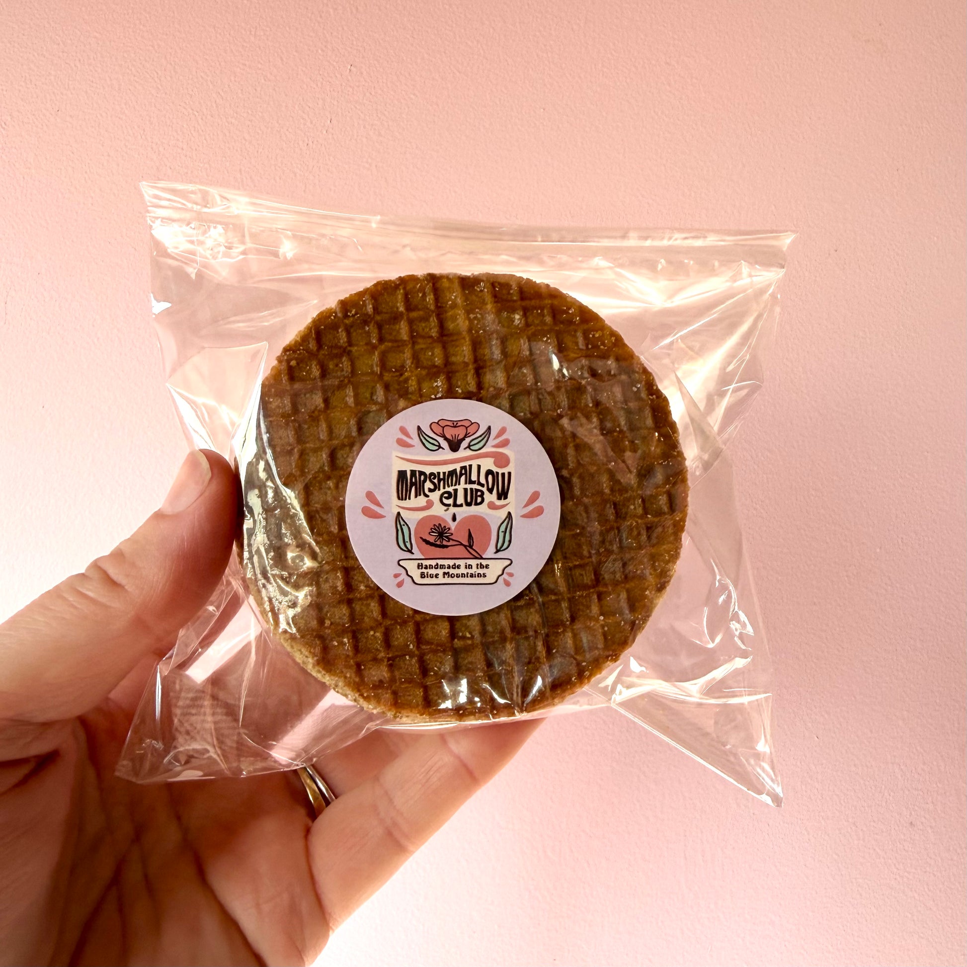 A hand holding a packaged round brown marshmallow waffle with a circular 'Marshmallow Club' sticker on a soft pink background.