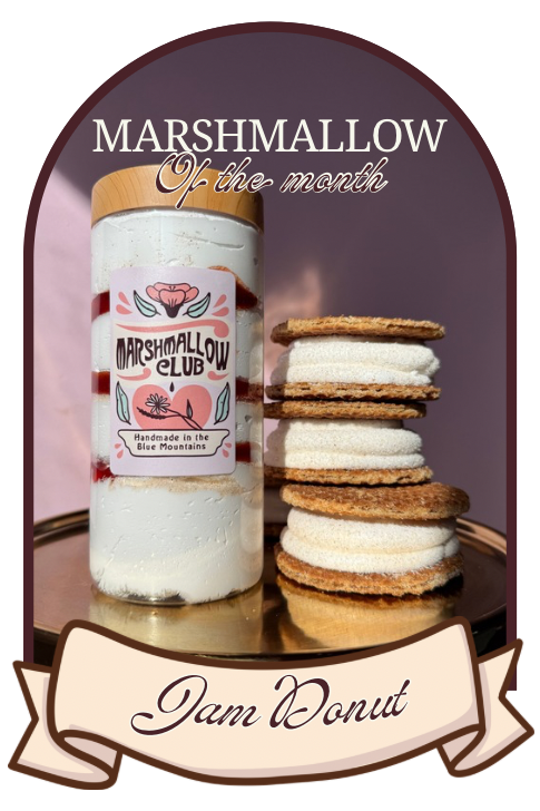 A promotional graphic for Marshmallow Club Marshmallow of the Month featuring a jar of white marshmallow cream with a wooden lid and a stack of marshmallow sandwiches on a gold tray, set inside an arched brown frame with text reading 'MARSHMALLOW of the month' and 'Jam Donut'.