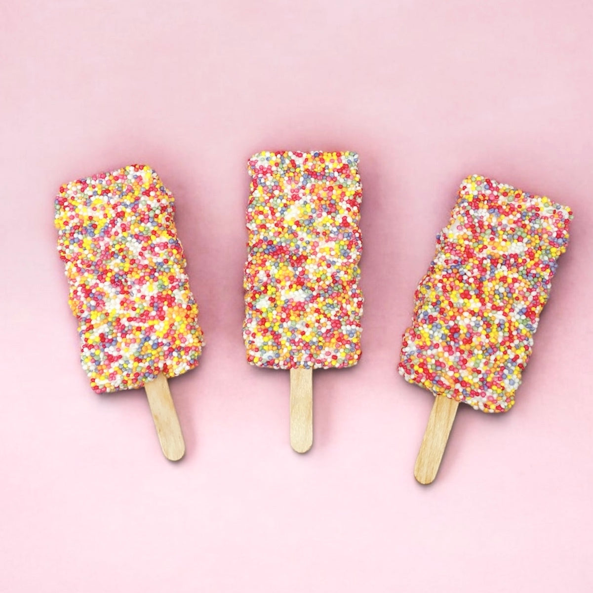Three colourful sprinkled marshmallow pops on a pink background